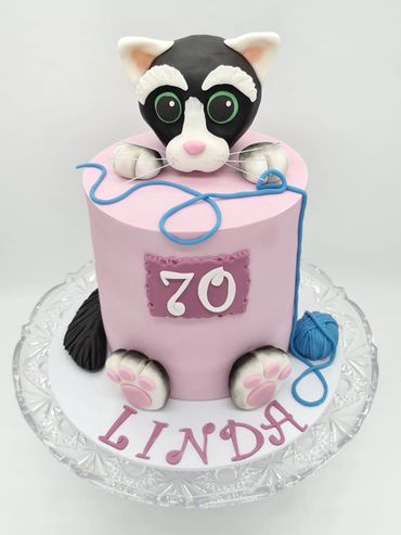 Cake covered in pink sugarpaste. Black and white cat popping out of the cake. Blue ball of yarn.