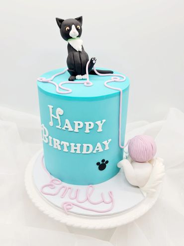 Blue sugarpaste cake with a black cat on top and a white cat playing with yarn at the bottom.