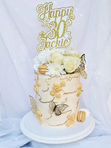 Cream cake covered in gold butterflies, macarons, sugarpaste roses. Fresh flowers and topper on top.