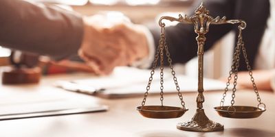 Scales of justice in focus with handshake blurred in background.