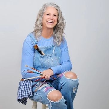 Smiling elderly woman with paintbrushes in overalls.