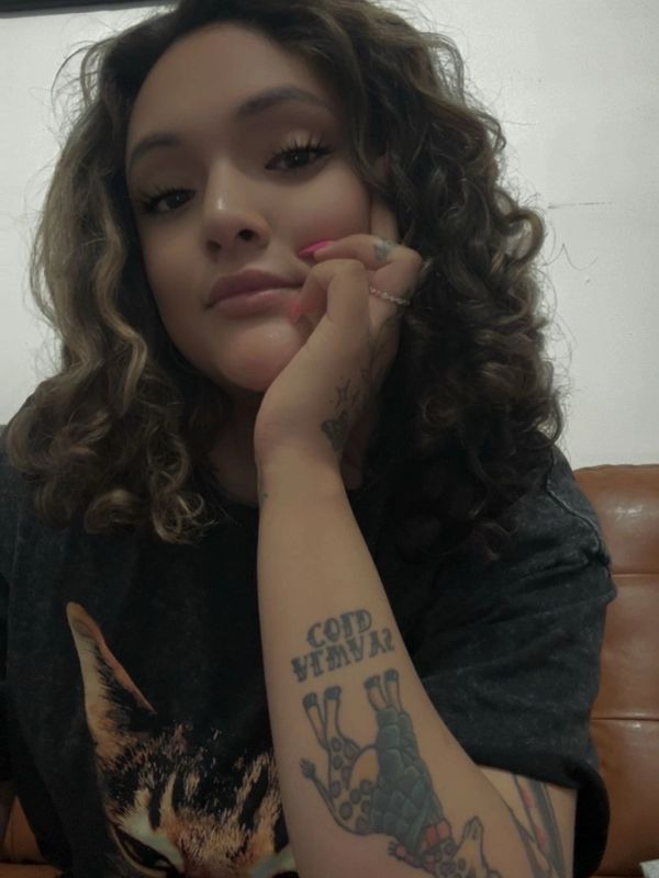 Woman with curly hair and a forearm tattoo, resting her face on her hand.