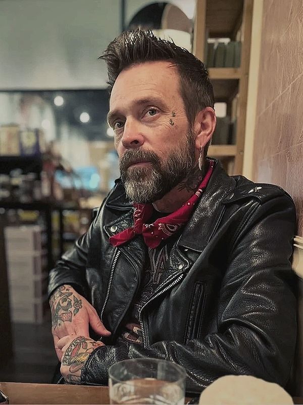 Man with tattoos, leather jacket, and red bandana at a table.