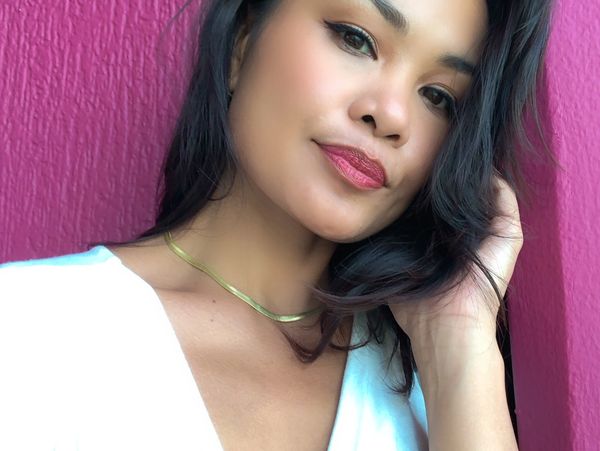 Woman with dark hair and gold necklace poses against a pink textured wall.
