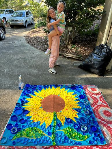 Two girls posing near a sunflower art made of bottle caps.