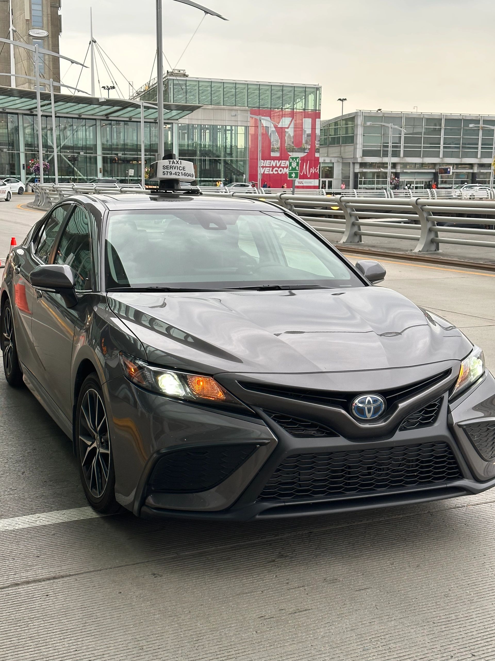 Gray Toyota taxi parked near a modern airport terminal on a cloudy day.