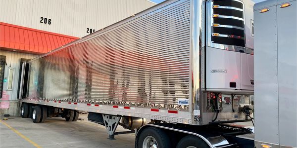 Shiny silver semi-trailer truck backed into loading dock under a blue sky.