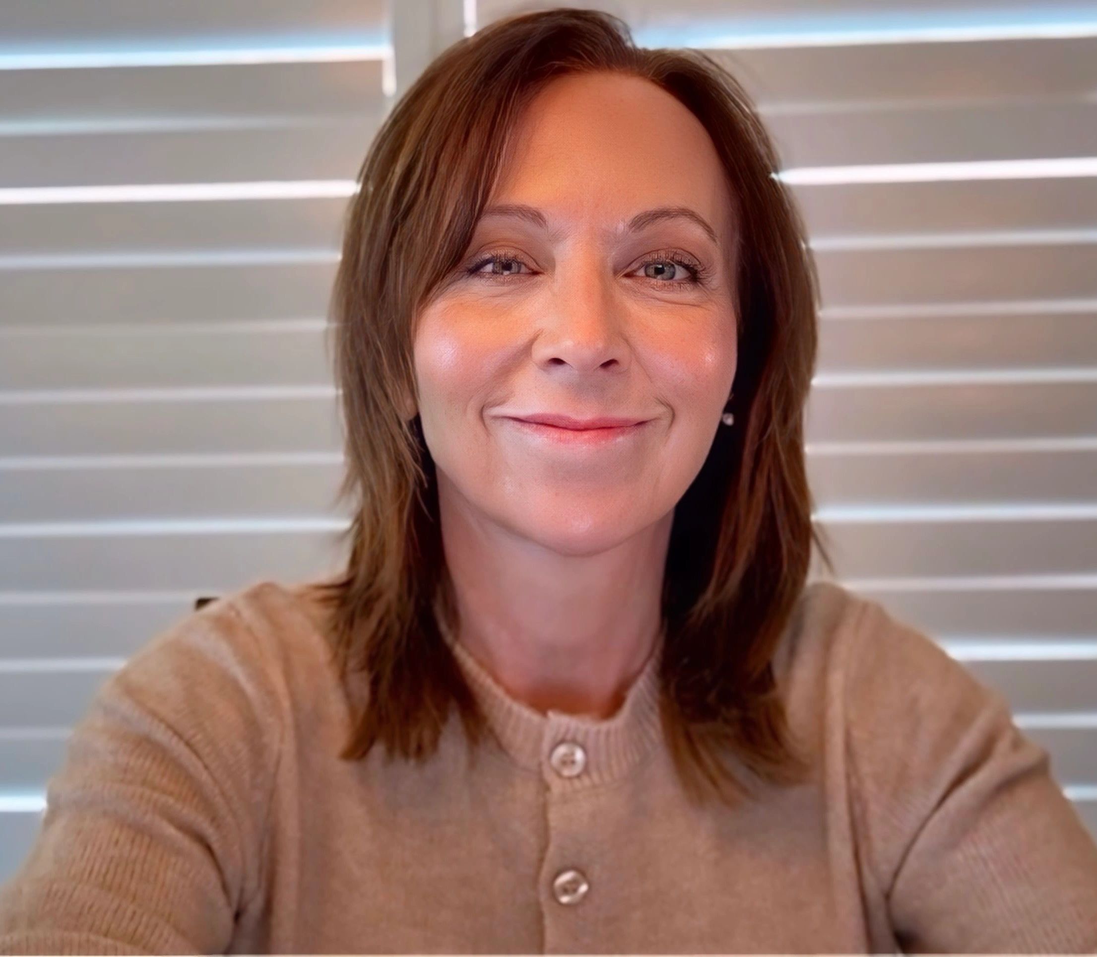 Smiling woman in beige sweater with shutter blinds background.