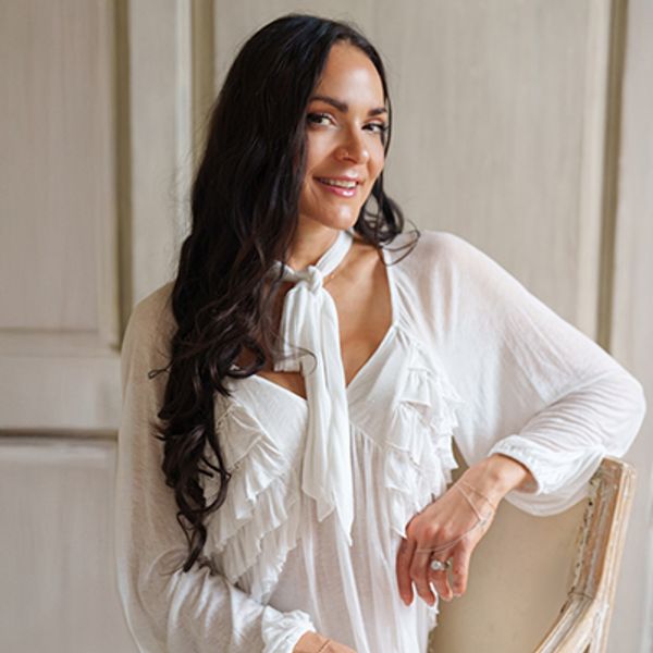 Smiling woman with long dark hair in a white blouse sitting on a chair.