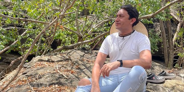 Man in white shirt and ripped jeans sitting barefoot on rocks surrounded by trees.