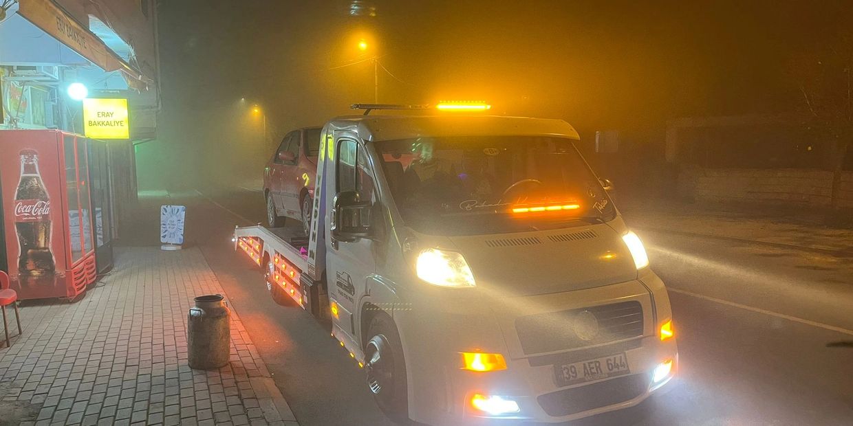 A tow truck carrying a red car at night with bright lights and fog.