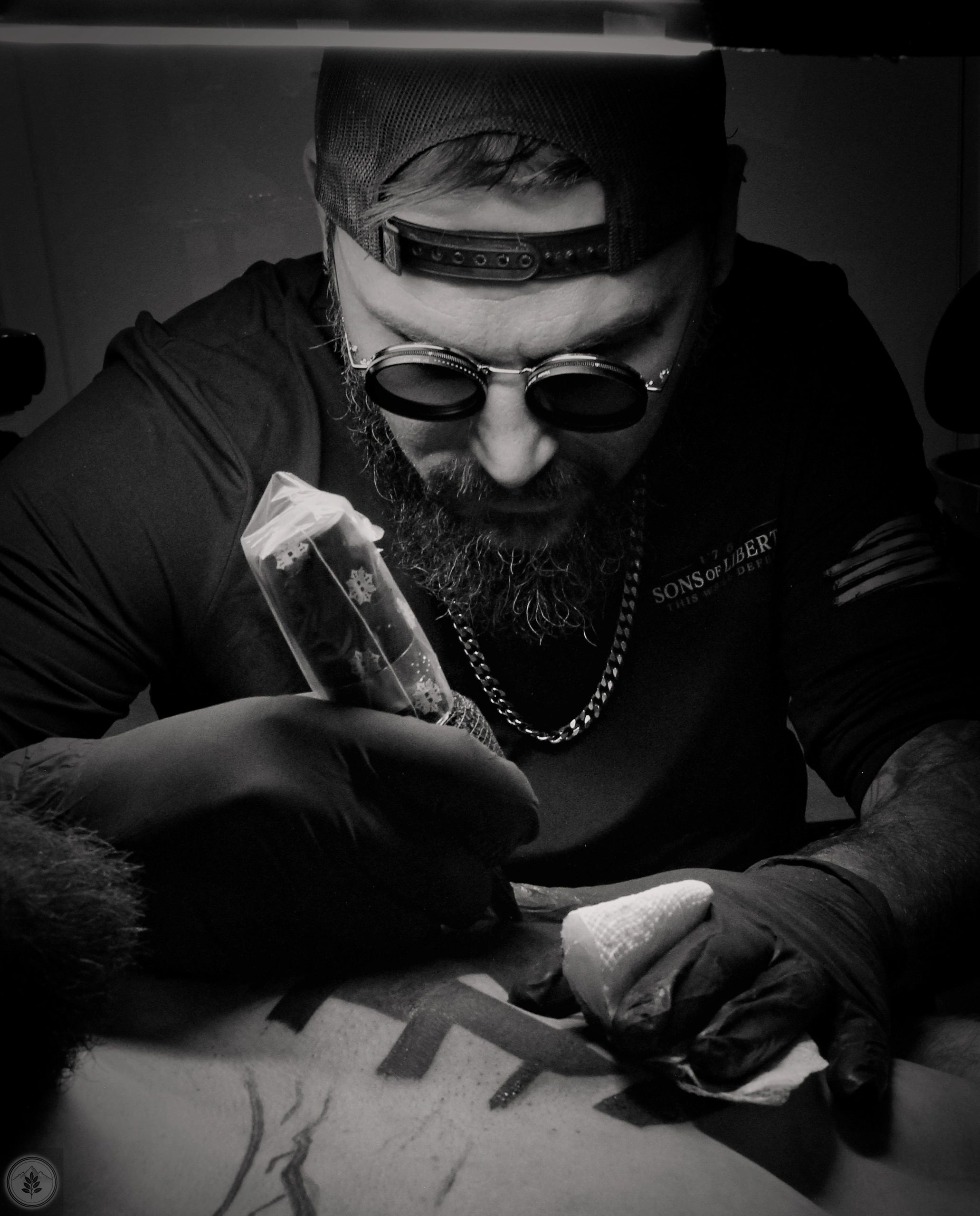 Man with glasses and gloves tattooing a person's chest in black and white.