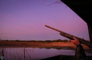 Person aiming shotgun near water at sunset.