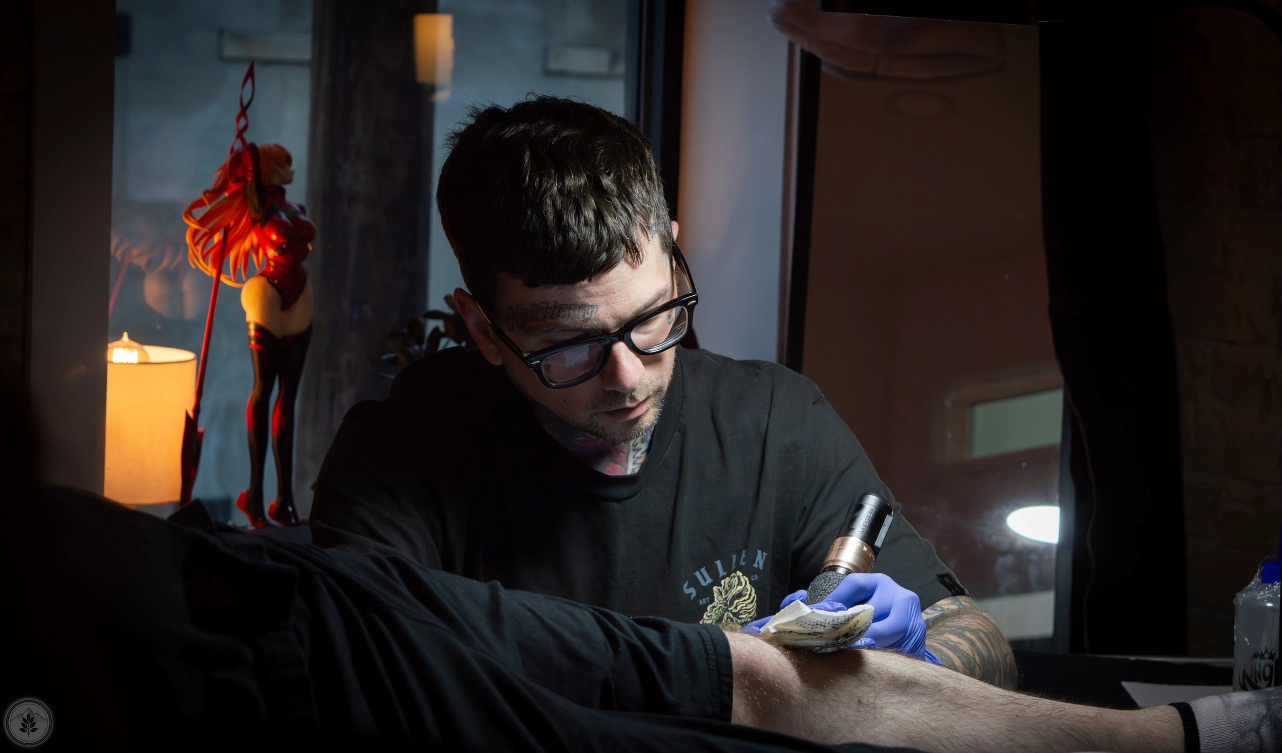 A tattoo artist concentrates while working on a client's leg tattoo in a dimly lit room.