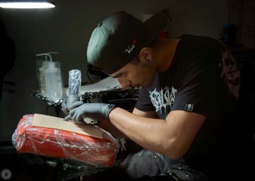 Man tattooing a client's arm in a dimly lit studio.