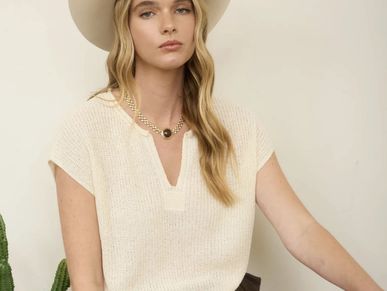 Young woman in a cream knit top and wide-brimmed hat poses indoors.