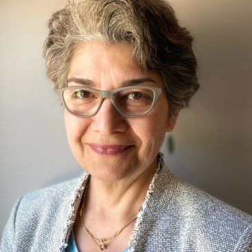 Smiling woman with gray hair and glasses wearing a blue top and textured jacket.
