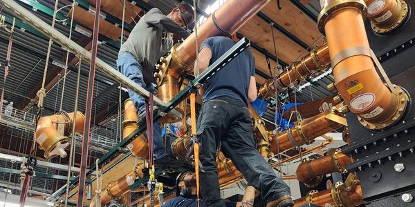 Workers install large copper pipes indoors.