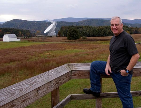Man posing by a wooden fence with mountains and a satellite dish in the background.