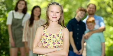 Smiling girl in floral dress stands confidently with family blurred in background outdoors.