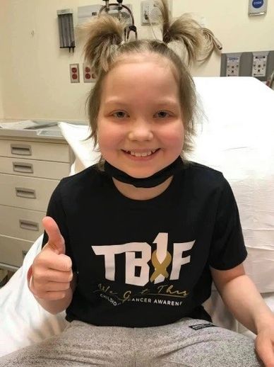 Smiling child with pigtails giving a thumbs up in a hospital room.