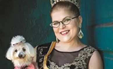 A woman wearing a crown and glasses holds a small dog dressed with a bow.