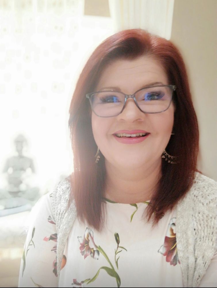 Smiling woman with glasses and red hair wearing a floral top and a knitted cardigan.
