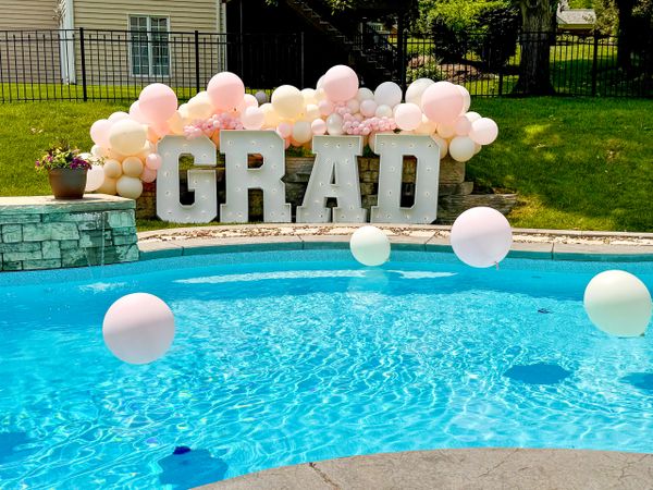 Bright GRAD letters with pastel balloons beside a clear swimming pool in a sunny backyard.