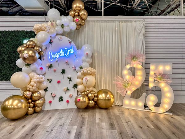 Elegant graduation backdrop with gold and white balloons, flowers, and illuminated '25' numbers.