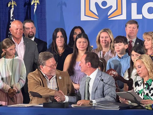 Two men shaking hands during a formal signing event surrounded by smiling people.
