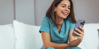 A nurse in scrubs smiling at her phone while sitting on a couch.