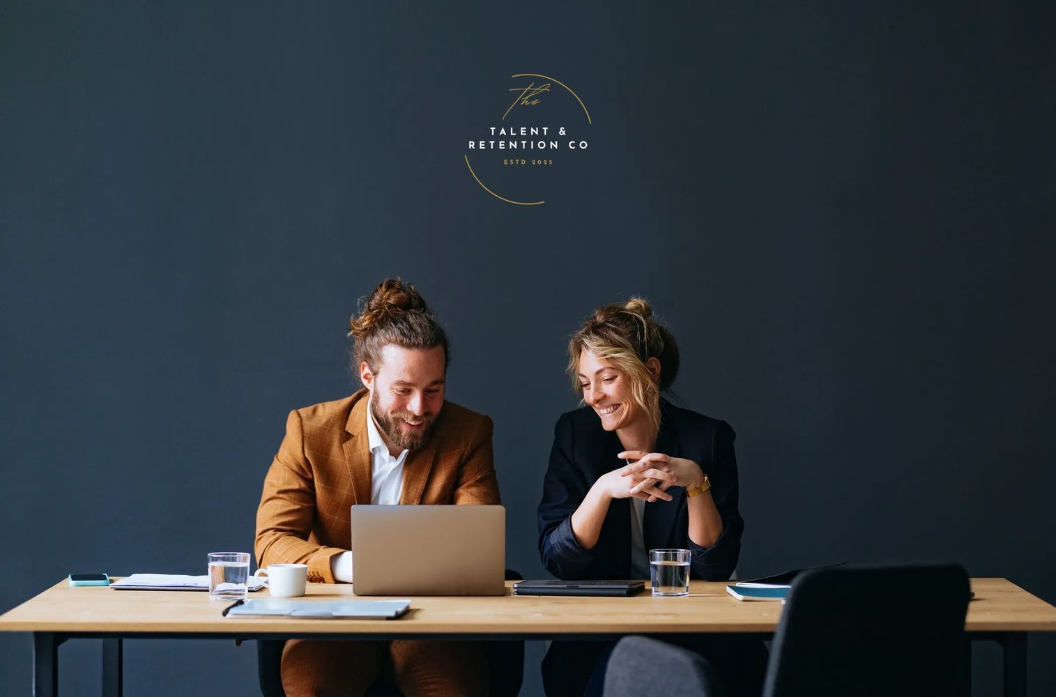 Two professionals smiling while working together at a laptop in a modern office.