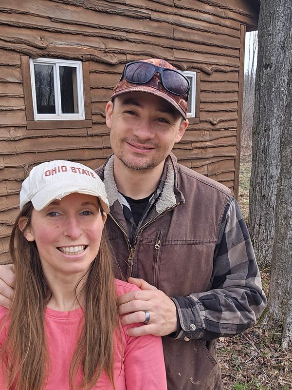 Smiling couple outdoors near a rustic wooden cabin in casual attire.