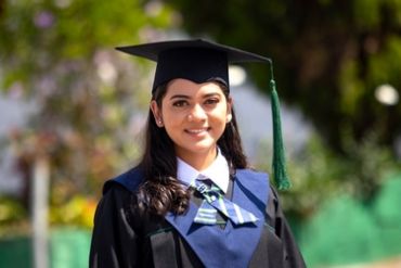 A young woman smiling in a graduation gown and cap outdoors.