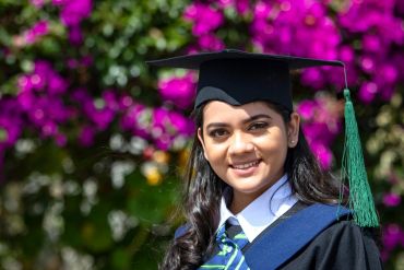 Smiling graduate woman in cap and gown with vibrant flowers behind.