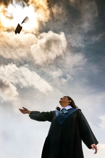 Graduate tossing cap into bright, cloudy sky, celebrating achievement.