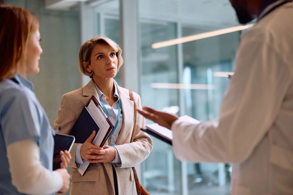 Medical professionals and a woman in a blazer discussing documents in a hospital corridor.
