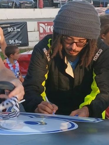 A man signs a race car hood while fans watch at a racetrack.