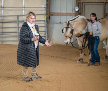 A woman in a black coat speaks while another holds a horse indoors.
