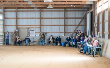 People seated along the walls inside a large indoor arena with dirt floor.