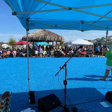 Outdoor stage setup with microphone and crowd under tents and a thatched roof.