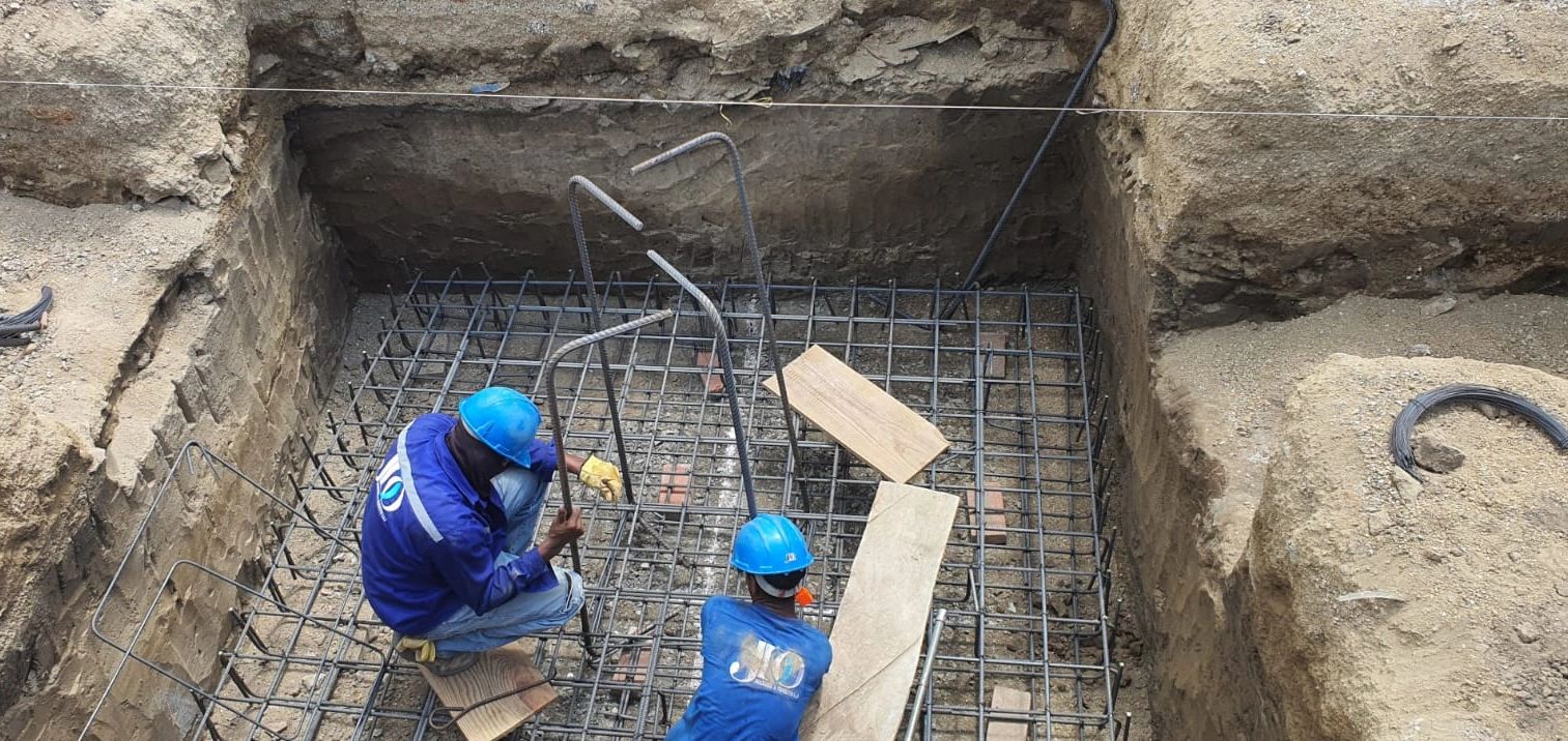 Two construction workers in blue helmets working on steel reinforcement in a foundation pit.