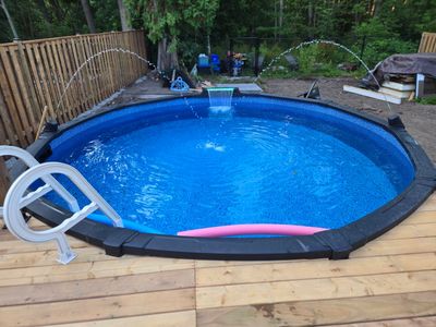 Above-ground pool with water fountains and wooden deck in a backyard.