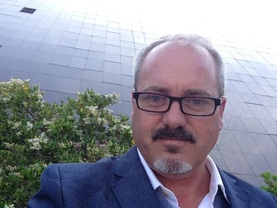 Man in glasses and suit outdoors near greenery and building.