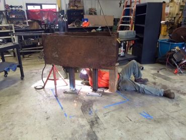 Person welding underneath a rusty metal structure in a workshop.