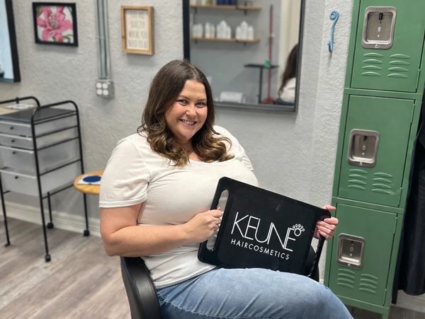 Woman sitting in salon chair holding a Keune Haircosmetics sign, smiling.
