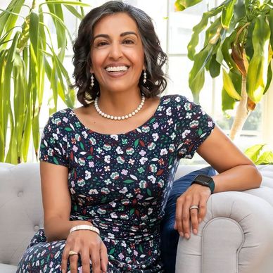 Smiling woman in floral dress sitting on a sofa surrounded by plants.
