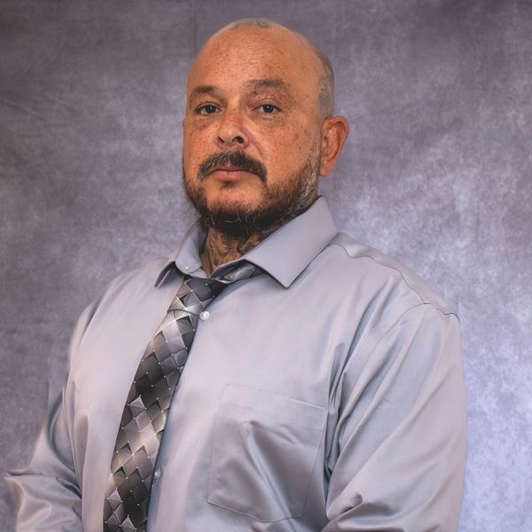 A serious man in a light gray shirt and patterned tie posing against a gray backdrop.