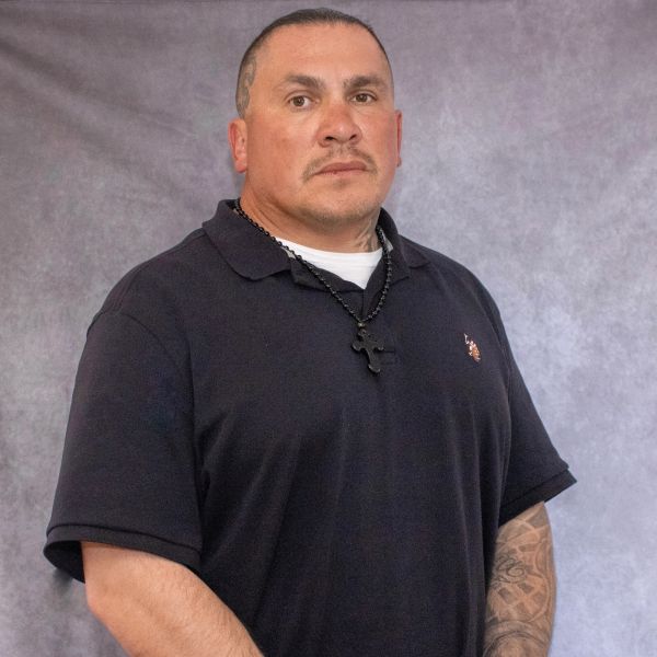 Man with tattooed arm wearing a black polo shirt and cross necklace.