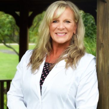 Smiling woman in a white coat standing outdoors by a wooden post.